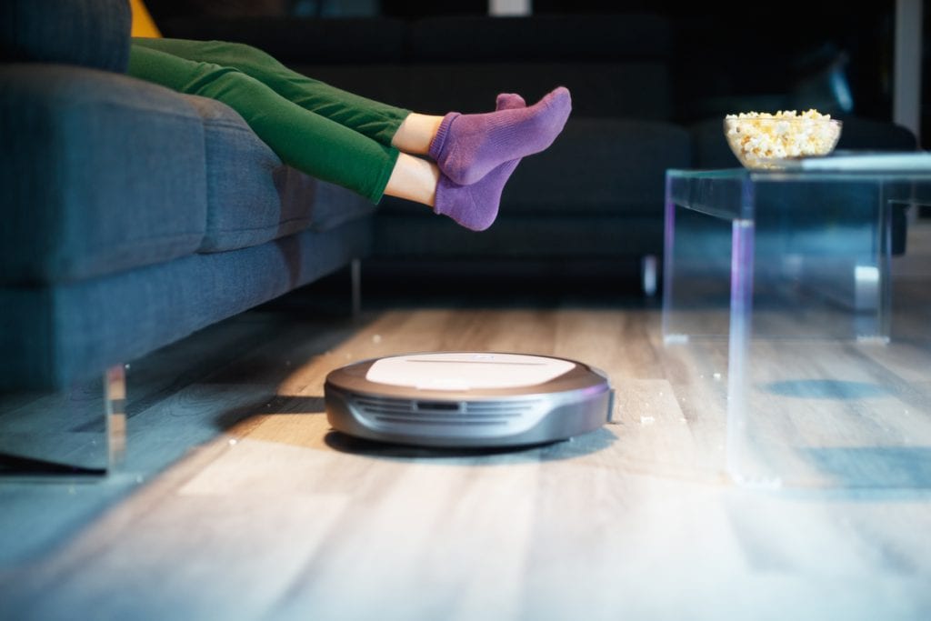 Young girl eating popcorn during movie night. Kid watches television on sofa. The child lifts feet up when a round robot vacuum cleaner passes to clean the dirty floor.